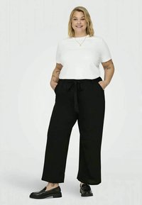 Crne široke hlače s vezanjem u struku, uparene s bijelim majicom kratkih rukava. Model nosi crne mokasine, pokazujući glatki, svakodnevni izgled.