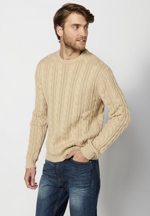 Beige kabelgebreide trui met een ronde halslijn en geribde manchet, gedragen met donkerblauwe jeans met lichte vervaging.