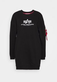 Robe noire à manches longues avec le logo et le texte Alpha Industries blancs sur la poitrine, avec une étiquette en tissu rouge attachée à la poche zippée sur la manche gauche.