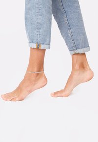 Sølvanklet på bar fod, med optrukne lysebl�å jeans. Foden er let fremadstillet, hvilket fremhæver krystal kædedesign og hudtekstur.