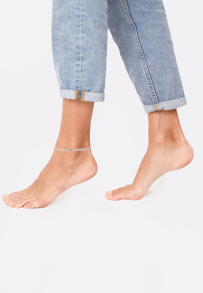 Sølvanklet på bar fod, med optrukne lyseblå jeans. Foden er let fremadstillet, hvilket fremhæver krystal kædedesign og hudtekstur.