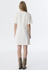 Kurzes cremefarbenes Kleid mit kurzen Ärmeln und geradem Schnitt, mit einer zentralen Naht auf dem Rücken. Kombiniert mit taupefarbenen Stiefeletten.
