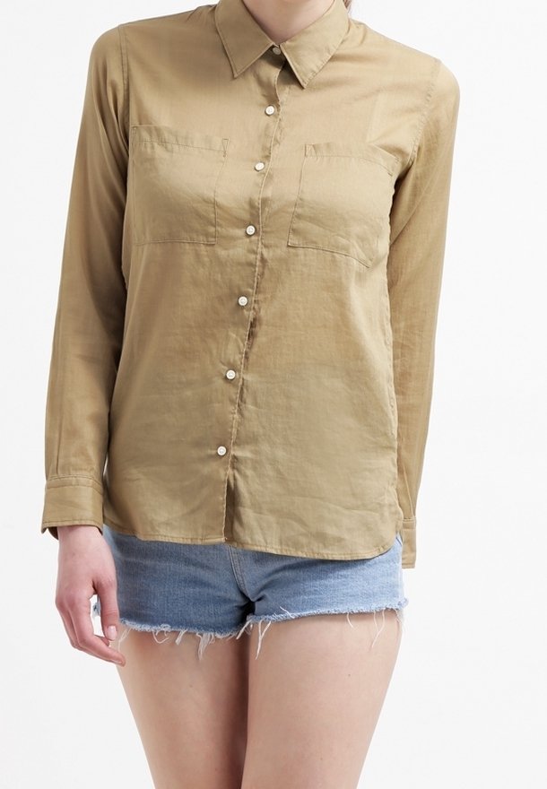 Chemise cintrée à manches longues couleur beige avec boutons, portée avec un short en denim bleu clair effiloché, sur une personne debout.