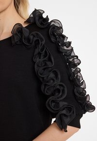 Schwarzes Oberteil mit gerüschten schwarzen Netzärmel im strukturierten Design. Der Stoff hat ein glattes Erscheinungsbild und eine figurbetonte Silhouette.