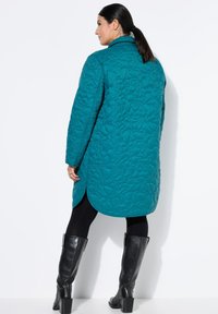 Teal kviltet lang frakke med et broderet blomsterdesign, sideslids og en notch-krave, parret med sorte knæhøje støvler.