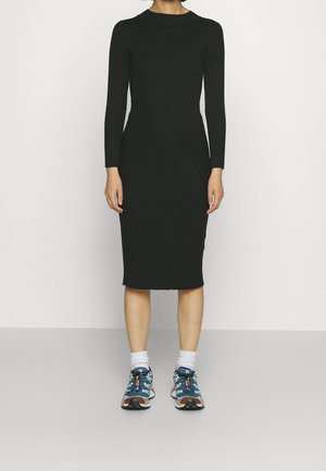 Persoon draagt een zwarte geribbelde midi-jurk met lange mouwen, gecombineerd met witte sokken en blauwe sneakers met patroon, staand tegen een effen achtergrond.