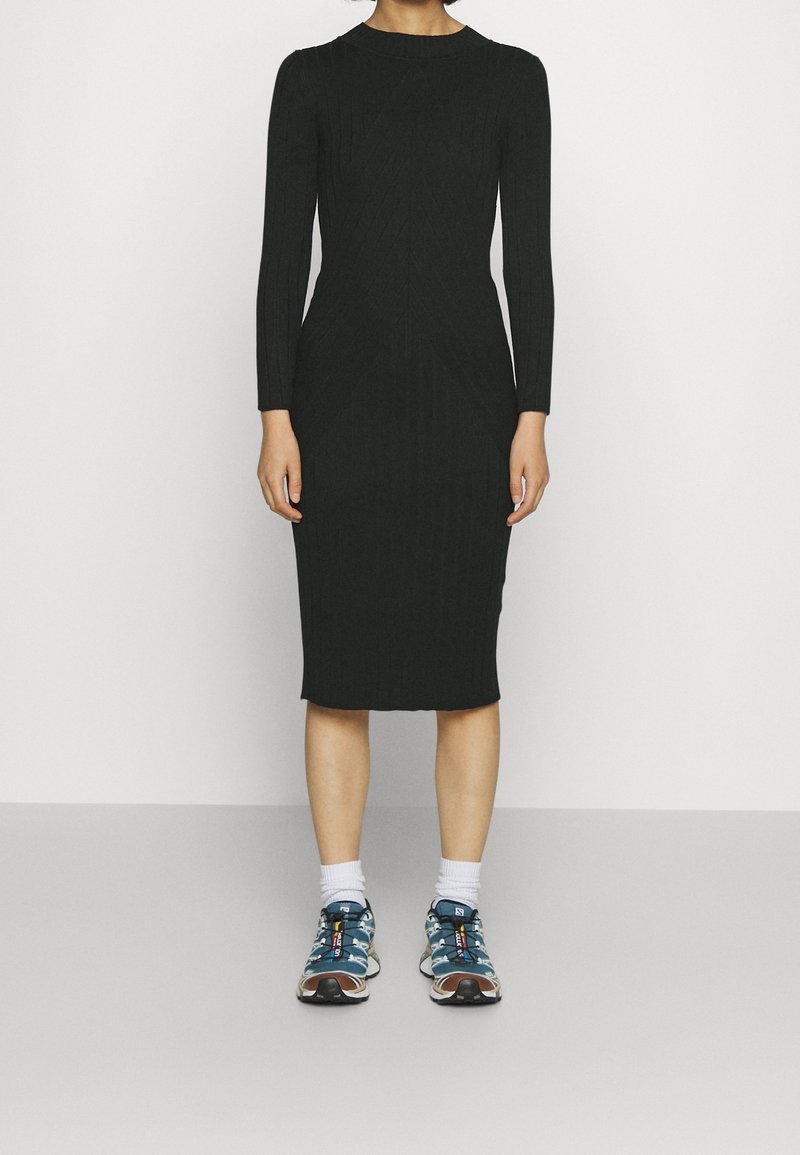 Personne portant une robe midi noire à manches longues côtelées, associée à des chaussettes blanches et des baskets bleues à motifs, debout devant un fond uni.