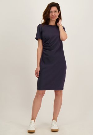 Vrouw met schouderlang bruin haar, dragend een marineblauwe jurk tot op de knie en witte sneakers, staand tegen een egale lichte achtergrond.