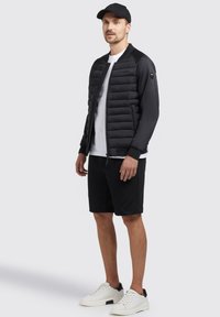Giubbotto bomber imbottito nero con tessuto liscio, t-shirt bianca sotto, pantaloni corti neri e sneakers bianche con dettagli neri.