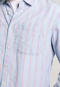 Vaaleansininen nappikauluspaita, jossa on pystysuorat punaiset ja valkoiset raidat. Rinnassa on tilkkutasku ja perinteinen kauluspaitakaulus.