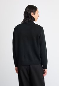 Juodas megztinis su aukštu kaklu ir griestu apačia. Turi ilgus rankovėmis ir laisvą siluetą, demonstruojantį minkštą tekstūrą.