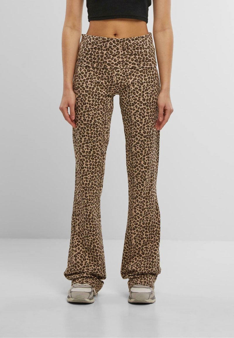 Femme portant un pantalon évasé taille haute à imprimé léopard marron et des baskets beiges, debout les mains détendues le long du corps sur un fond gris.