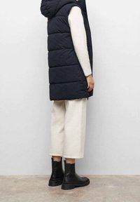 Tmavě modrá péřová vesta s kapucí, zdobená prošívanými částmi. Kombinována s béžovými širokými kalhotami a černými kotníkovými botami.