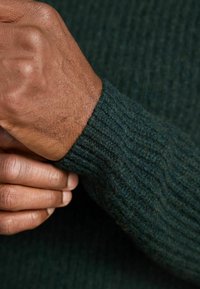 Dunkelgrüner gestrickter Pulloverärmel mit gerippter Struktur. Eine Hand hält die Manschette und zeigt die Dicke und Weichheit des Stoffes.