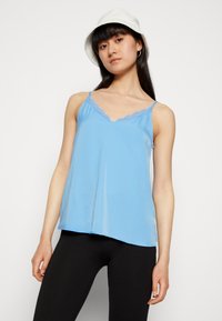 Camisola sem mangas de um azul claro, com renda ao longo do decote, feita de um tecido suave, combinada com leggings pretas e um chapéu branco tipo bucket.