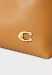 Oda tan spalvos odinė rankinė su lygiu paviršiumi, išsiskirianti išryškintu auksinės spalvos "C" logotipu, kaip akcento detalė.