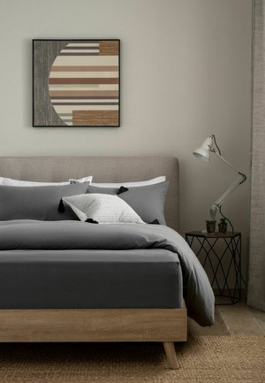 Bett mit grauer Bettwäsche und weißen und grauen Kissen, Holzrahmen, geometrische Wandkunst darüber und Beistelltisch mit Schreibtischlampe und kleinen Pflanzen.