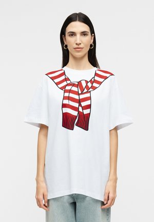 Γυναίκα που φοράει λευκό φαρδύ t-shirt με γραφικό κόκκινου και λευκού ριγέ πουλόβερ δεμένο στον λαιμό, συνδυασμένο με ανοιχτόχρωμο τζιν.