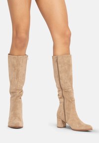Botas de ante en color beige hasta la rodilla, con tacón apilado y cremallera lateral. Suave textura y un diseño holgado realzan la silueta general.