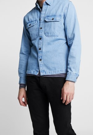 Personne portant une chemise en jean bleu clair avec des poches poitrine boutonnées par-dessus un t-shirt à manches longues rayé et un pantalon noir sur un fond uni.