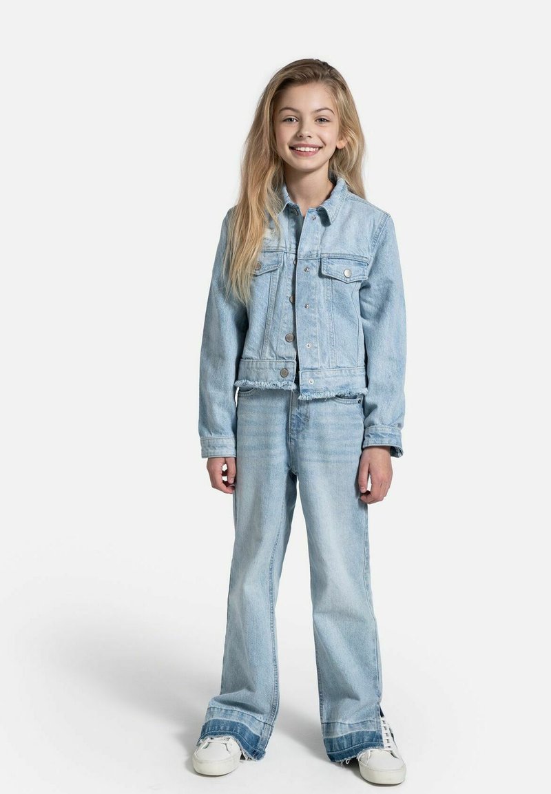 Jasnoniebieska kurtka dżinsowa z kieszeniami z przodu i postrzępionym dołem, w połączeniu z dopasowanymi jasnoniebieskimi dżinsami dzwonami i białymi trampkami.