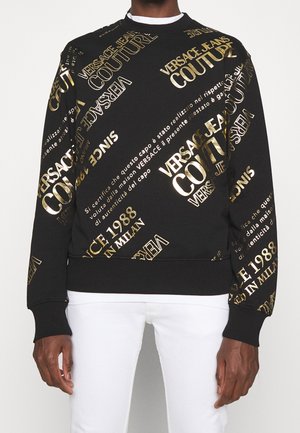 Sweat noir avec grand texte doré "Versace Jeans Couture" et texte italien doré supplémentaire, porté avec un pantalon blanc sur une personne.