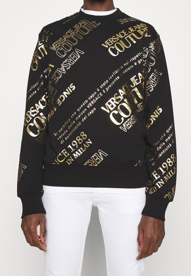Sweat noir avec grand texte doré "Versace Jeans Couture" et texte italien doré supplémentaire, porté avec un pantalon blanc sur une personne.