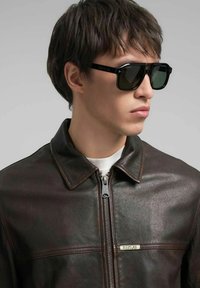 Giacca in pelle marrone scuro con chiusura a zip, caratterizzata da un colletto e da un dettaglio con logo metallico. Occhiali da sole neri con lenti verdi.