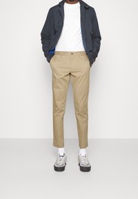 Kevyet tummansininen takki, jossa on vetoketjukiinnitys ja sininen vuori, yhdistettynä beigeihin chinokalsareihin ja harmaisiin lenkkikenkiin, joissa on mustat pohjat.