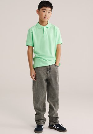 Zelené polokošeľa a sivé voľné rifle s jemnou textúrou a uvoľneným strihom. Oblečenie je doplnené tmavými teniskami s bielymi pruhmi.