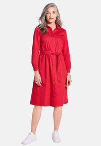 Rotes Hemdblusenkleid mit langen Ärmeln und tailliertem Schnitt. Aus Baumwolle, knielang, mit Kragen und Taschen. Kombiniert mit weißen Sneakern.