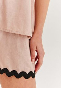 Svetloružový top s vrstvovým dizajnom, s čiernym scalloped lemovaním na okraji. Tkanina vyzerá mäkko a textúrovane. Ruka spočíva na okraji.