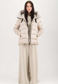 Beige Daunenjacke mit großem Kapuzen, gestepptem Design, Frontreißverschluss und schwarzen Akzenten. Getragen über einem passenden, hellbeigen Outfit.