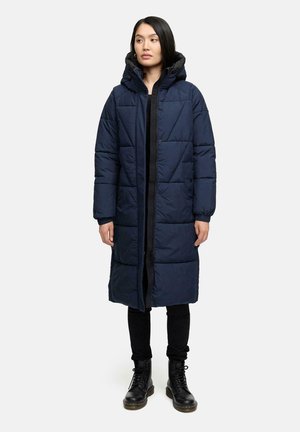Marineblauer gepolsterter Mantel mit Kapuze, der über ein geometrisches Steppmuster, eine Knopfverschlus und schwarze Akzente verfügt. Kombiniert mit schwarzen Hosen und Stiefeln.