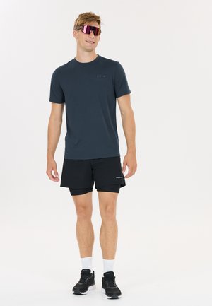 Man draagt een donker sportshirt en korte broek, zwarte hardloopschoenen, witte sokken en reflecterende sportzonnebril, staand tegen een witte achtergrond.