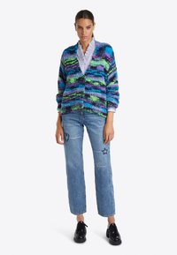 Cardigan en tricot multicolore avec des rayures bleues, vertes et violettes ; coupe décontractée avec des fermetures à boutons ; associé à un jean bleu clair et des chaussures noires.