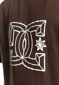 Camiseta de algodón marrón con diseño geométrico bordado en blanco y acento de estrella. El diseño incluye formas entrelazadas y contornos.