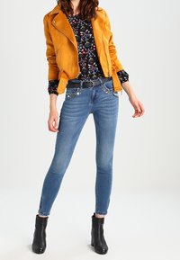 Veste motard en daim jaune, haut à manches longues à motifs floraux, jean skinny bleu orné de décorations, et bottines noires. Textures lisses, coupe ajustée.
