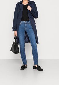 Laivastonsininen sadetakki, musta t-paita, siniset farkut ja mustat loafersit. Pitäen pientä mustaa käsilaukkua. Sileät pinnat, klassisia muotoiluelementtejä.