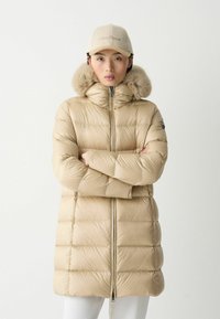 Bežs puffer parka ar mākslīgās vilnas kapuci, ar rāvējslēdzēja aizdari, horizontālu quiltu rakstu un ribota adījuma cimdiem. Cepure ar izšūtu logo.