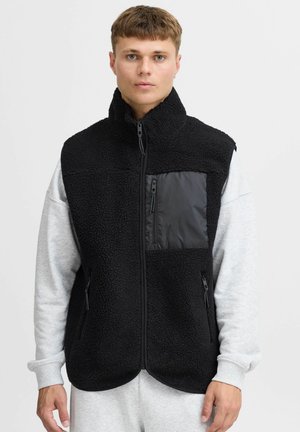 Sort fleece vest med høj krave, en frontlomme med sort stof og sideloger, båret over en lysegrå sweatshirts.