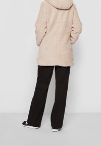 Beige faux fur jakke med hætte, der har en afslappet pasform og tekstureret overflade, parret med sorte cargo bukser og atletiske sko.