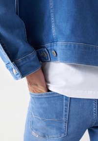 Osoba oblečená v modré džínové bundě a džínách, s pravou rukou v zadní kapse, pod bundou viditelná bílá košile.