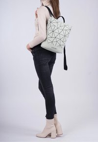 Le sac à dos présente un extérieur gris clair avec un motif géométrique, des accents noirs et une poignée supérieure robuste. Des bretelles noires s'étendent des côtés.