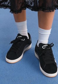 Black suede sneakers with white rubber soles, featuring black laces and a circular logo on the side. Worn with white ribbed socks.