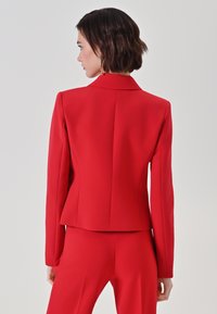 Blazer rosso con silhouette aderente, rever a incavo, maniche lunghe e texture liscia. Presenta cuciture sartoriali e un retro strutturato.