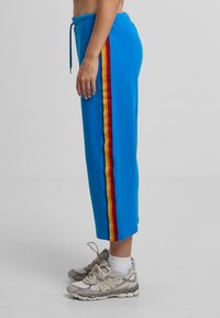 Modré tepláky se žlutým a červeným bočním pruhem, elastickým páskem a volným, zkráceným střihem. Nosené s béžovými teniskami.