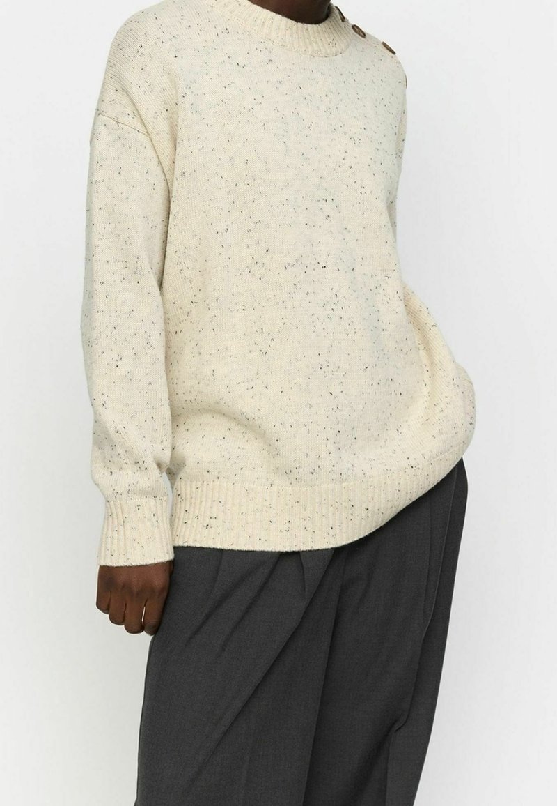 Personne portant un pull en tricot crème moucheté avec des détails de boutons sur l'épaule et un pantalon plissé gris foncé, debout avec la main dans la poche.