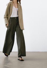 Beige oversized skjorte med en enkelt brystlomme, parret med viddebenede mørkegrønne bukser og sorte thong-sandaler på en lys baggrund.