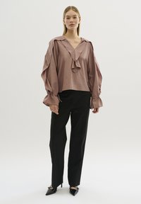 Luftige, mauve Bluse mit V-Ausschnitt, geschichteten Rüschendetails und voluminösen Ärmeln. Kombiniert mit schwarzen, taillierten Hosen und spitzen Absätzen.